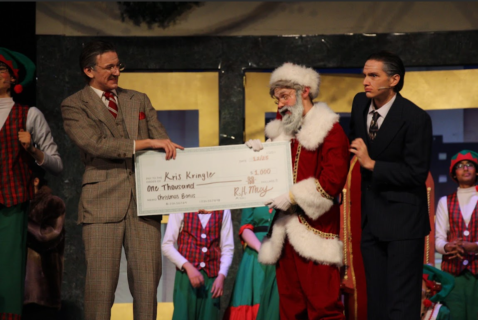 Justin Malec ('26) as Kris Kringle onstage, accepting his Christmas bonus from AJ Gangewere ('25) as Macy and Sam Becker ('25) as Bloomingdale.