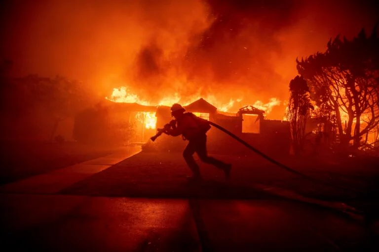 A firefighter scrambles to battle the Palisades Fire as it burns down a home in the Pacific Palisades neighborhood  of Los Angeles (Courtesy of AP Photo/Ethan Swope).